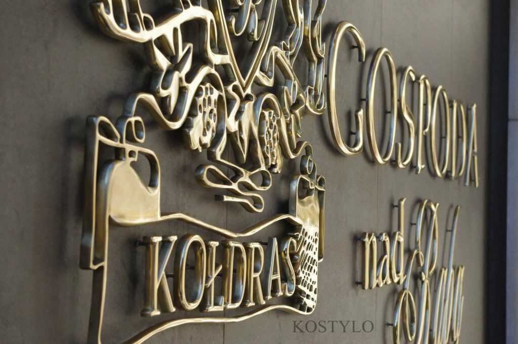 an angled close-up of beautiful openwork logo in the form of bas-relief, handcrafted from brass polished to high gloss in Kostylo atelier